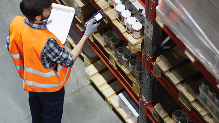 A warehouse worker in a safety vest checks inventory with a handheld scanner and clipboard, standing next to shelves stocked with boxes and containers.