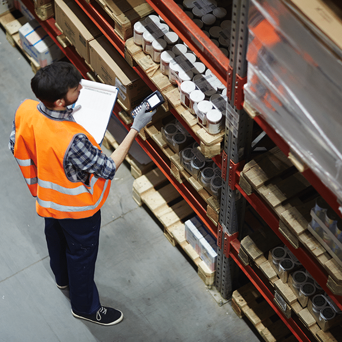 A warehouse worker in a safety vest checks inventory with a handheld scanner and clipboard, standing next to shelves stocked with boxes and containers.