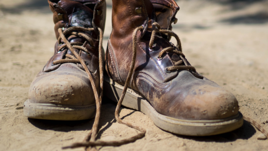 A pair of worn, brown leather work boots with untied laces are placed on sandy ground in sunlight, showing scuff marks and signs of heavy use.