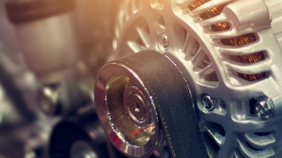 Close-up of a car engine showing a belt wrapped around a metal pulley and an alternator, with mechanical components blurred in the background. Warm lighting highlights the metal surfaces.