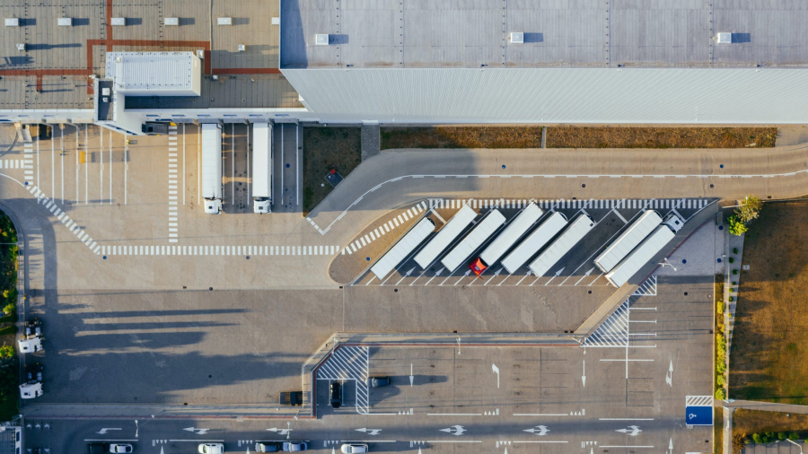 Aerial view of a large warehouse with multiple loading docks, several semi-trailers parked in a row, and a parking lot with cars in the lower section of the image.