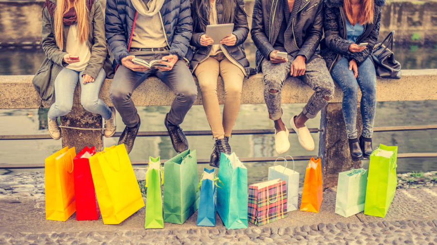 Five people sit on a stone railing by the water, facing forward, holding phones or books. Colorful shopping bags are lined up on the ground in front of them. Only their lower bodies are visible.
