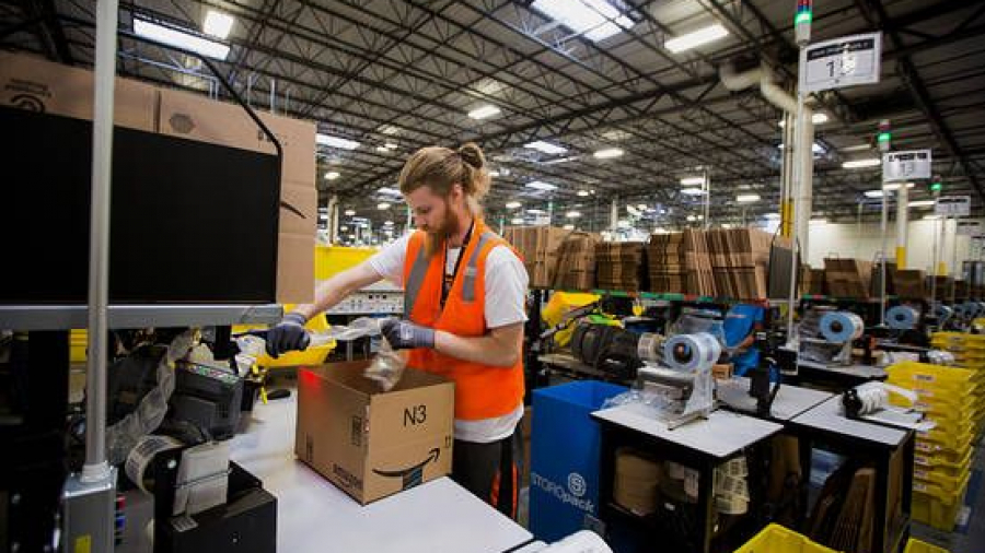 A warehouse worker in an orange safety vest packs items into a cardboard box at a workstation in a large, brightly lit industrial facility filled with equipment and stacks of boxes.