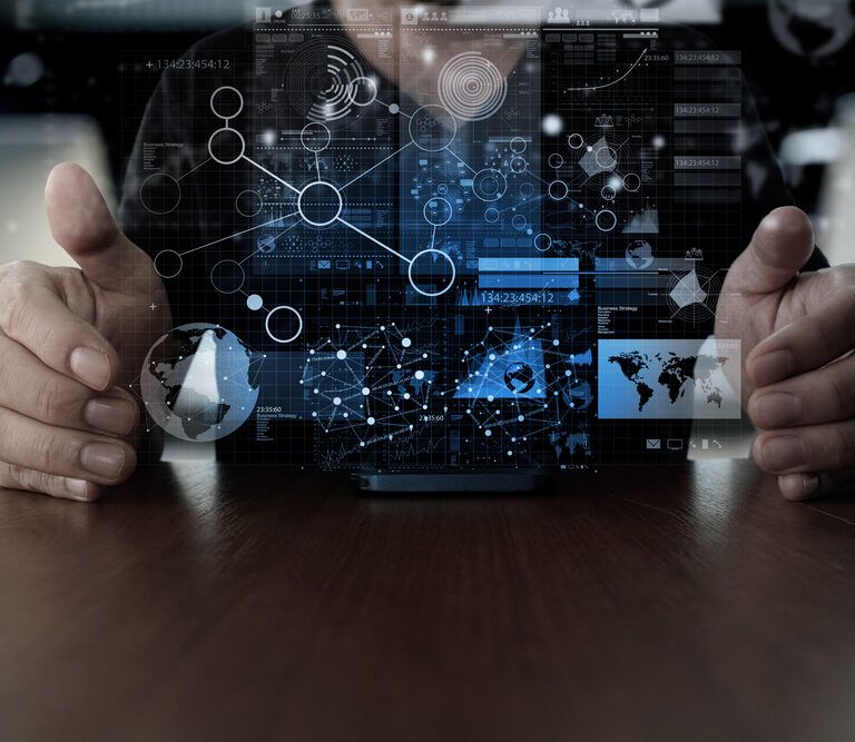 A person’s hands are hovering around a digital interface with floating futuristic graphics, including charts, maps, and network connections, projected above a desk. The scene suggests technology, data analysis, or innovation.