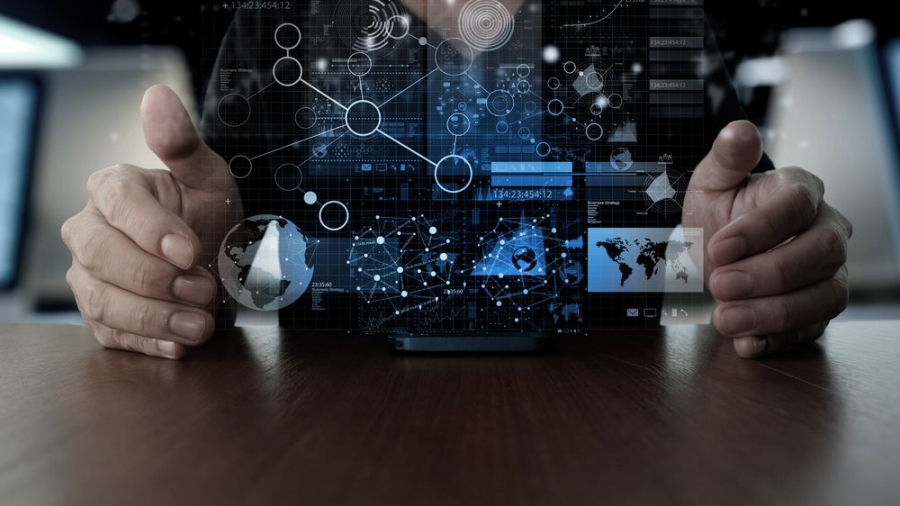 A person’s hands are hovering around a digital interface with floating futuristic graphics, including charts, maps, and network connections, projected above a desk. The scene suggests technology, data analysis, or innovation.