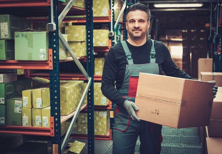 A man in work overalls and gloves stands in a warehouse, holding a large cardboard box. Shelves filled with various packed boxes are visible behind him. The environment is well-lit and organized.