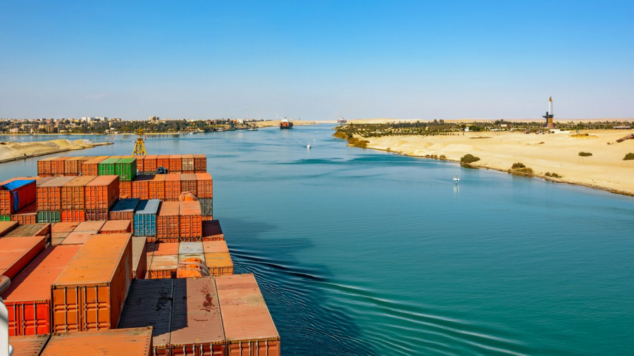 A large cargo ship with stacked shipping containers sails through the Suez Canal, bordered by sandy desert on the right and buildings in the distance under a clear blue sky.