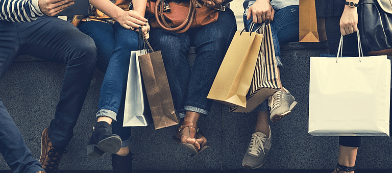 Four people sit on a bench holding various shopping bags. Only their legs, hands, and bags are visible. They wear casual clothing and different types of shoes, suggesting a shopping outing.