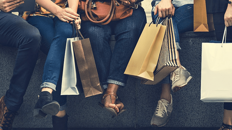 Four people sit on a bench holding various shopping bags. Only their legs, hands, and bags are visible. They wear casual clothing and different types of shoes, suggesting a shopping outing.