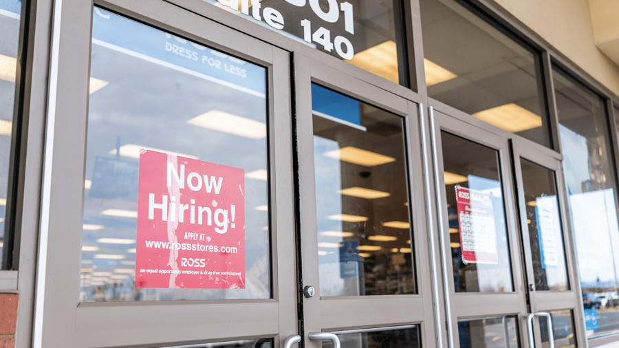 Glass doors of a retail store with a prominent red Now Hiring! sign displayed, reflecting the parking lot outside; store lights and aisles are visible through the glass.