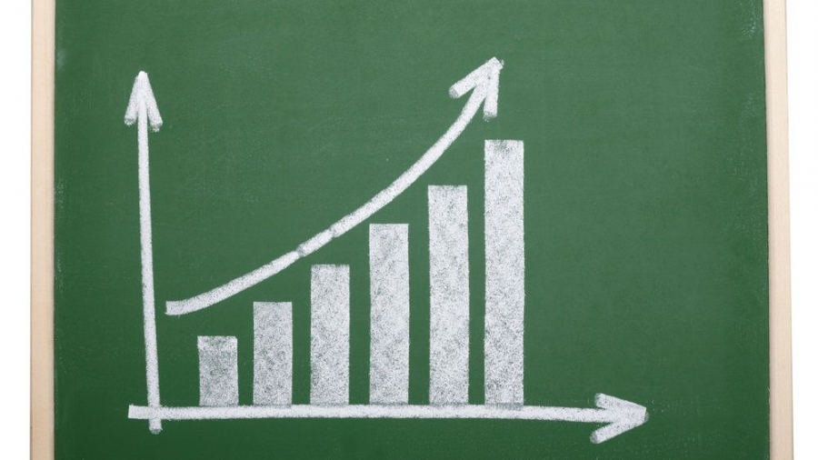 A green chalkboard with a hand-drawn bar graph in white chalk, showing increasing bars from left to right and an upward curving arrow above the bars, indicating growth or progress.