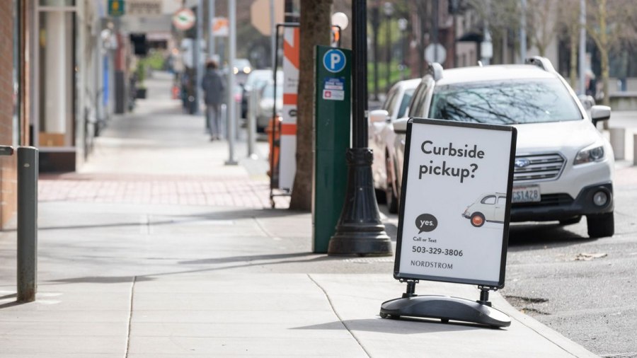 A sidewalk sign advertises curbside pickup for Nordstrom, featuring a graphic of a car and a phone number, with parked cars and people walking in the background on a city street.