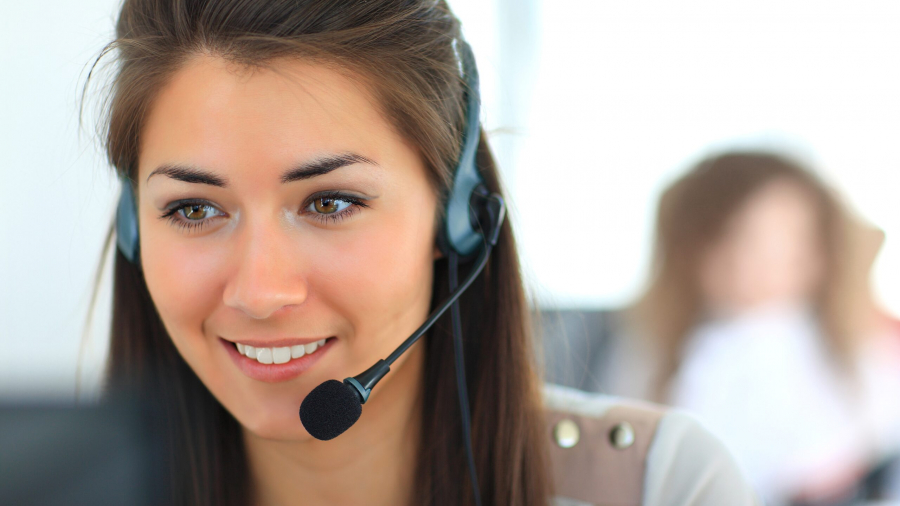 A woman wearing a headset with a microphone is smiling while looking at a computer screen, suggesting she is working in a customer service or call center environment. The background is blurred.