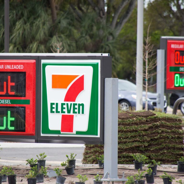 A 7-Eleven gas station sign displays Out for both regular unleaded and diesel fuels, indicating they are unavailable. Another similar sign is visible in the background. Small plants surround the signs.