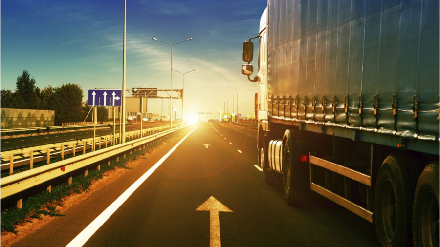 A large truck drives down a highway at sunrise or sunset, with bright sunlight ahead, road signs on the left, and a clear sky overhead. The road features lane markings and guardrails.