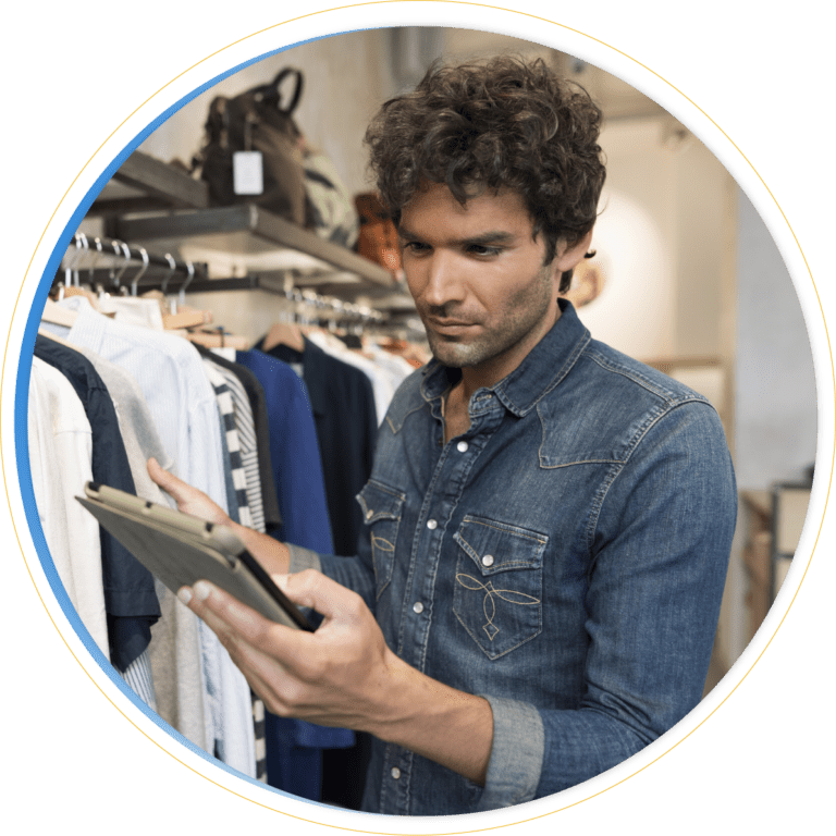 A person wearing a denim shirt stands in a clothing store, reviewing merchandising strategy on a tablet while holding it with one hand. Racks of clothes and bags are visible in the background.