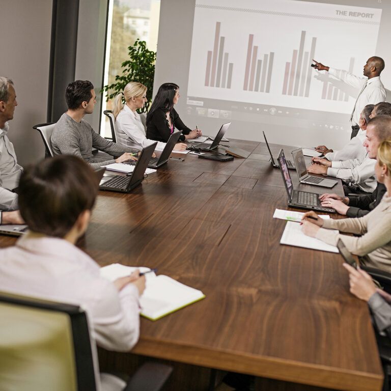 A group of people in business attire sit around a conference table with laptops and notepads, as a presenter stands near a screen displaying a bar graph during a Microsoft Dynamics AX upgrade meeting.