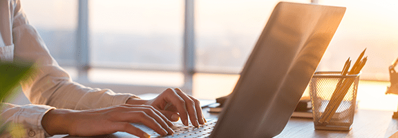 A person types on a laptop at a desk with sunlight streaming through large windows. A pencil holder with pens and pencils is visible beside the laptop.