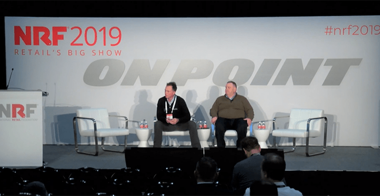 Two men sit on a panel stage at NRF 2019 Retail’s Big Show, with the words “ON POINT” in large letters behind them and empty chairs beside them. The audience is partially visible in the foreground.