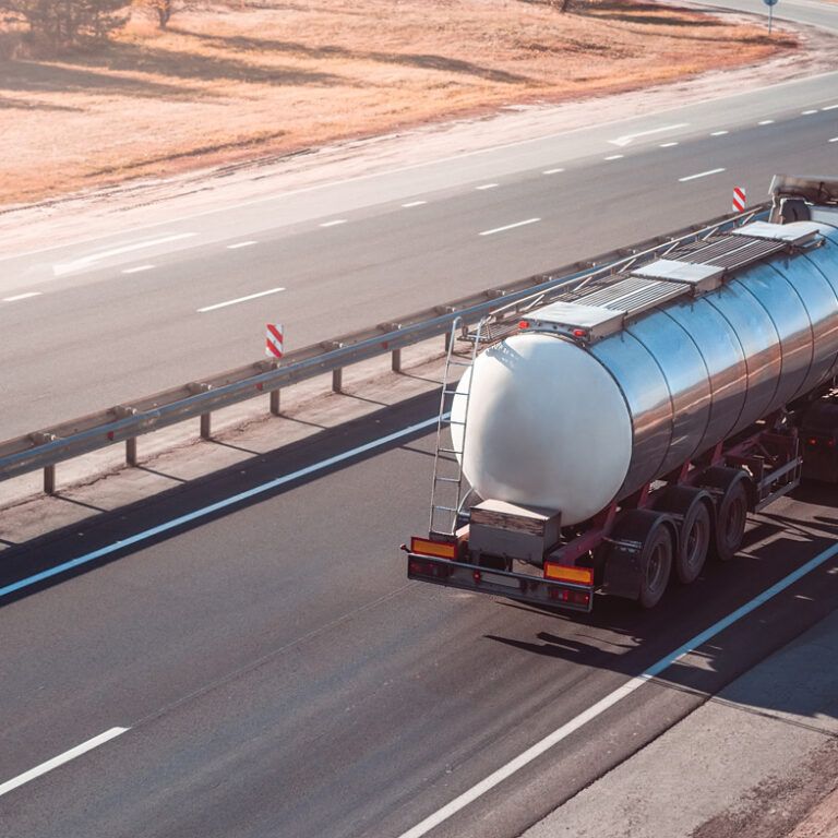 A white tanker truck drives on a mostly empty multi-lane highway bordered by grass and guardrails, with clear weather and gentle sunlight.