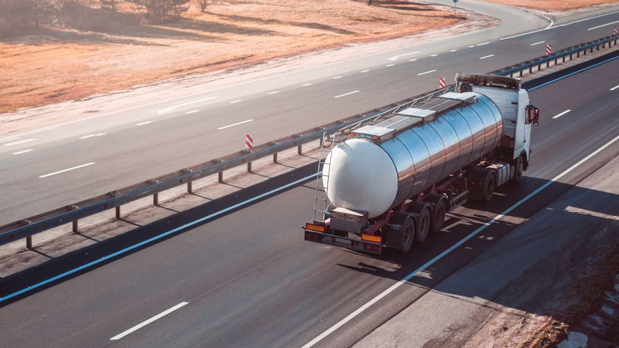 A white tanker truck drives on a mostly empty multi-lane highway bordered by grass and guardrails, with clear weather and gentle sunlight.