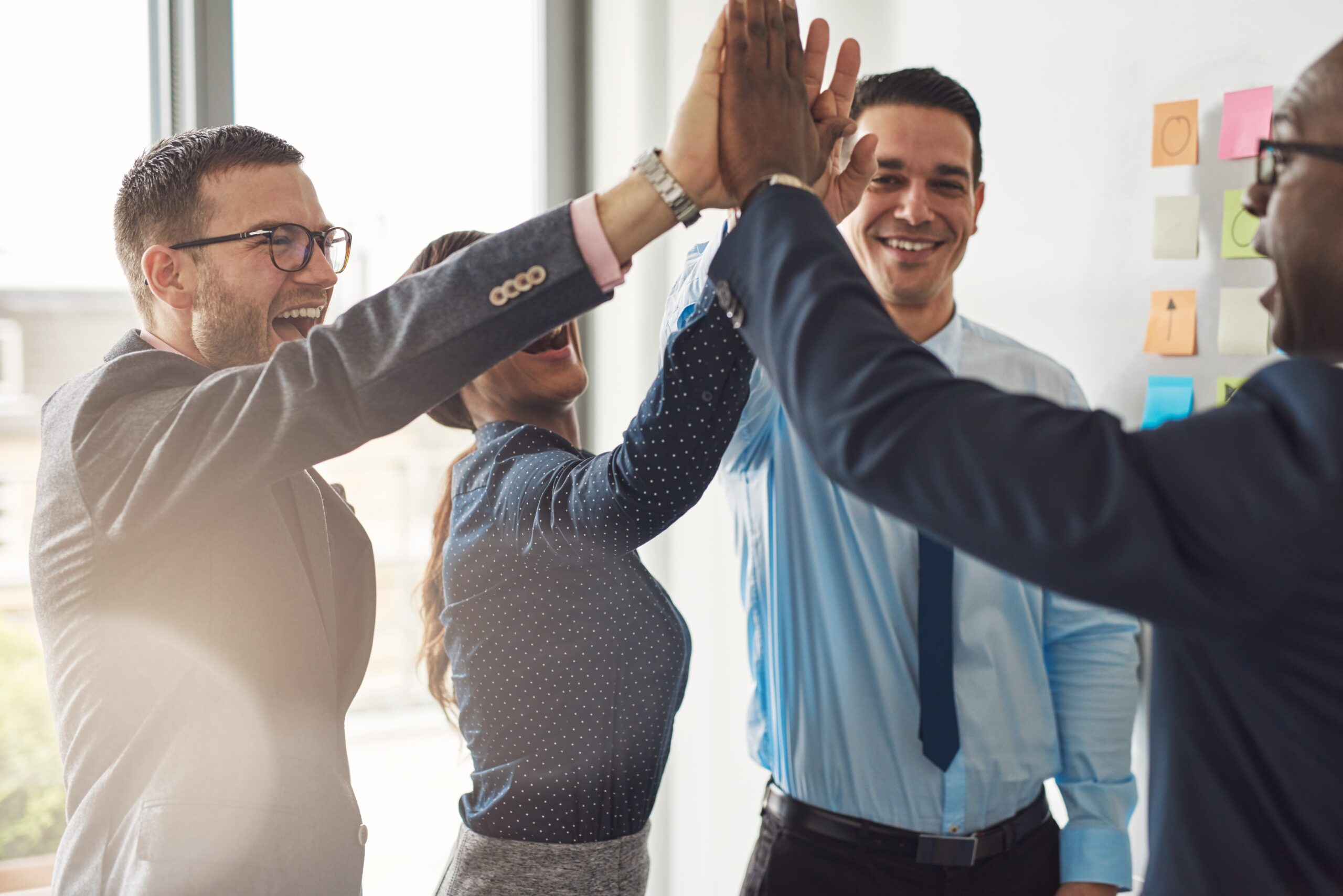 Four professionally dressed people stand in an office, smiling and giving each other high fives. Colorful sticky notes are visible on the wall behind them, suggesting a collaborative, successful team meeting.