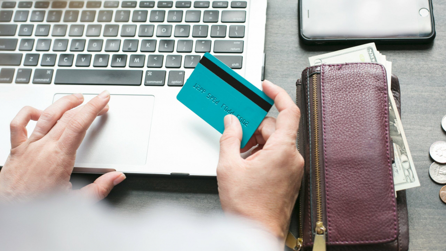 A person holding a blue credit card sits at a laptop, with a purple wallet, cash, coins, and a smartphone on the table nearby. The scene suggests online shopping or banking.
