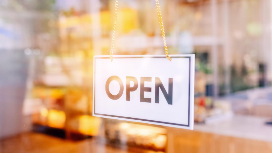 A white rectangular sign with the word “OPEN” in bold black letters hangs on a glass door, with a blurred interior of a shop or café visible in the background.
