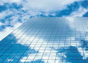A tall glass building reflects a blue sky with white clouds, creating an almost seamless blend between the architecture and the sky above.