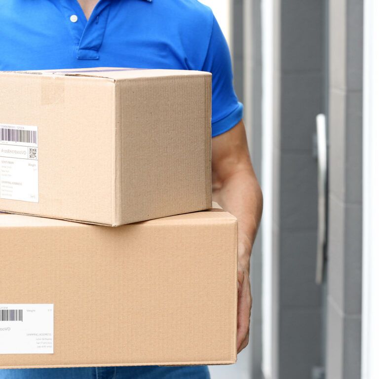 A person in a blue shirt holds two cardboard delivery boxes, standing outside a building with gray walls and a door. Both boxes have shipping labels attached.