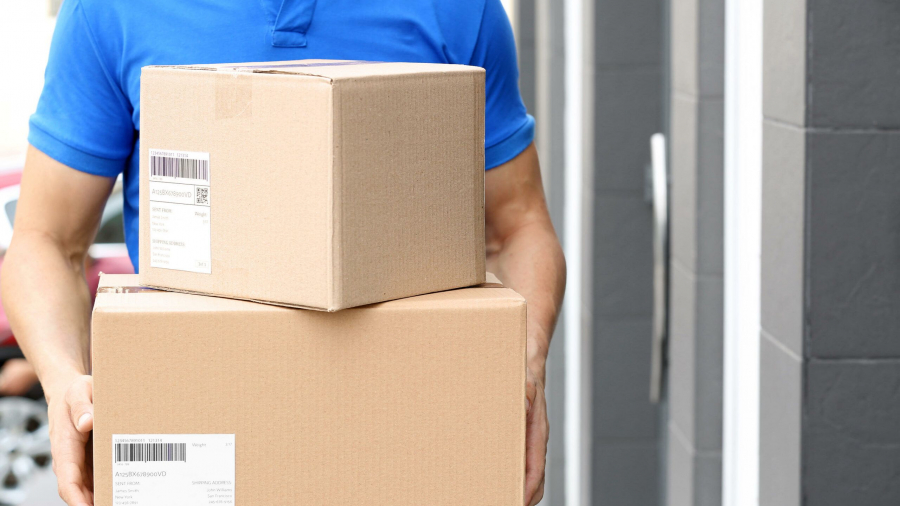 A person in a blue shirt holds two cardboard delivery boxes, standing outside a building with gray walls and a door. Both boxes have shipping labels attached.