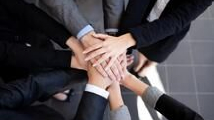A group of people wearing business attire stand in a circle and stack their hands together in the center, symbolizing teamwork and unity.