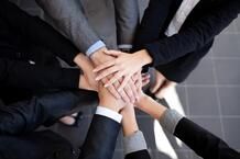 A group of people wearing business attire stand in a circle and stack their hands together in the center, symbolizing teamwork and unity.