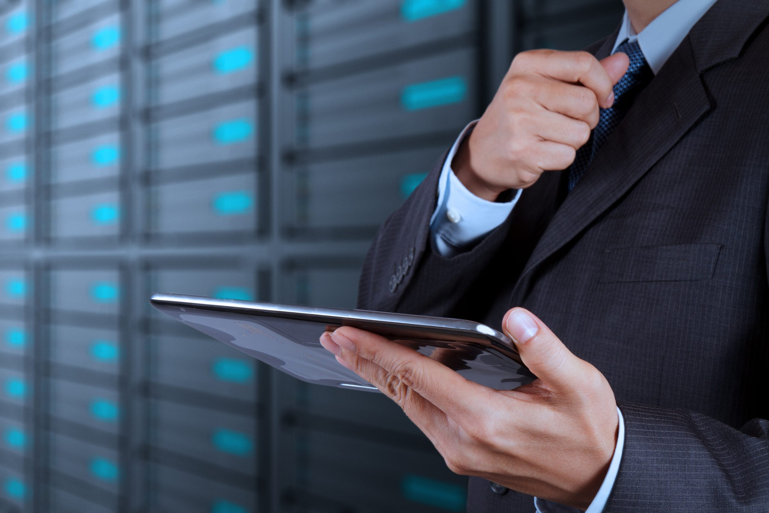 A person in a suit holds a tablet and adjusts their tie, standing in front of blurred server racks in a data center.