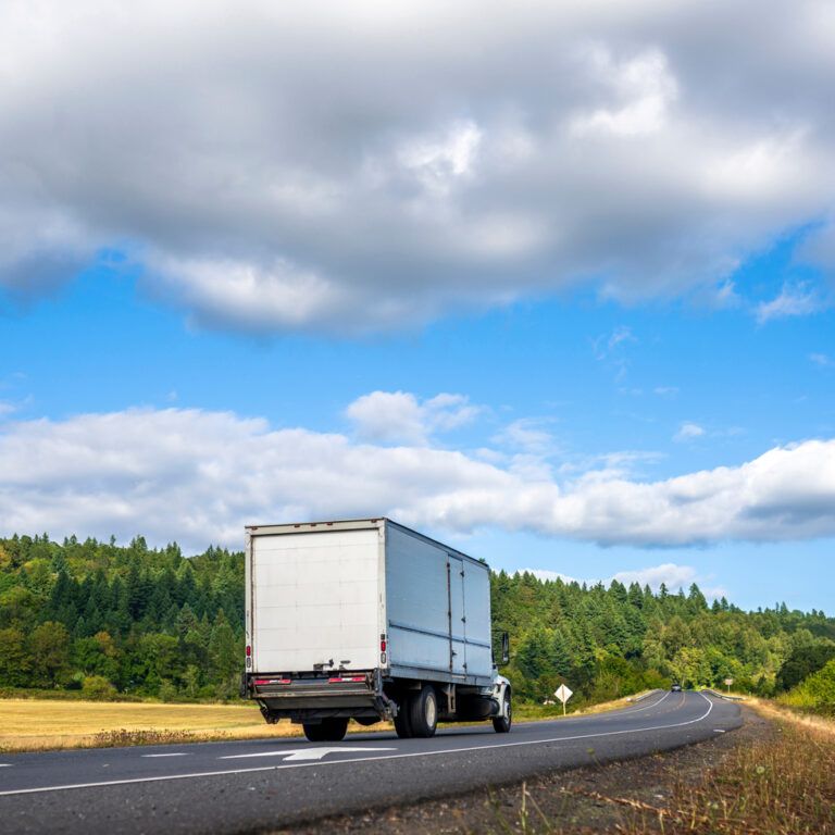 A white semi-truck drives down a curving rural road bordered by green trees and fields under a partly cloudy blue sky.