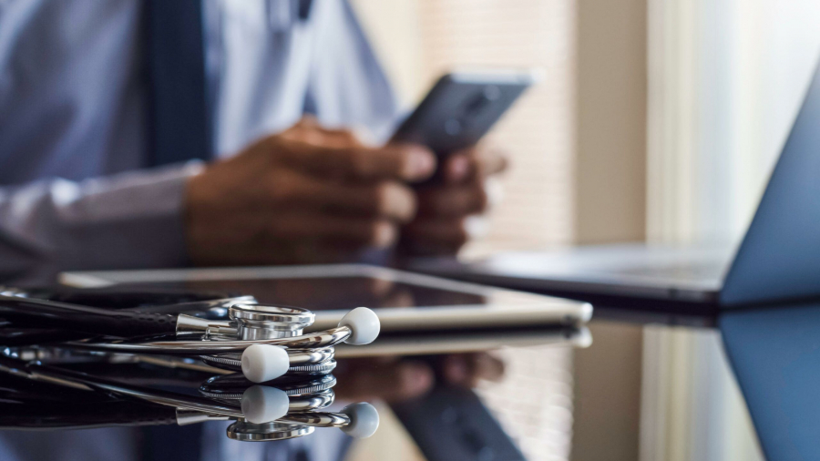 A stethoscope rests on a desk near a tablet and laptop, while a person in business attire uses a smartphone in the background.