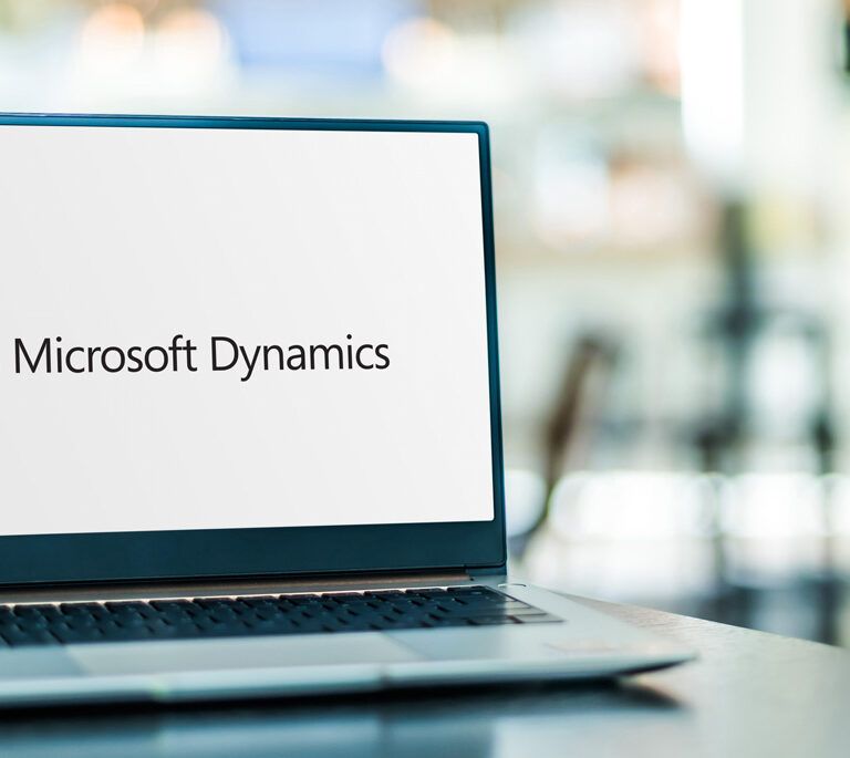 A laptop on a table displays the Microsoft Dynamics logo on its screen, with a blurred background of an office or cafe setting.