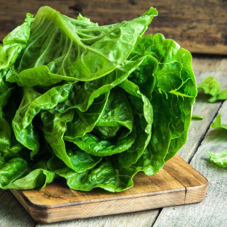 A head of fresh green lettuce sits on a wooden cutting board placed on a rustic wooden table, with a few loose leaves scattered around.