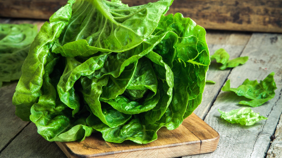 A head of fresh green lettuce sits on a wooden cutting board placed on a rustic wooden table, with a few loose leaves scattered around.