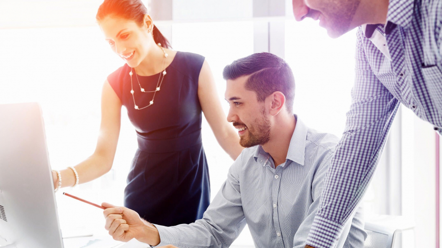 Three colleagues, two men and one woman, collaborate at a desk in a bright office, smiling and looking at a computer screen while discussing work. The woman is pointing at the monitor.