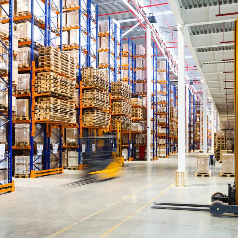 Large warehouse with tall shelves stacked with boxes, showcasing efficient slotting for organized storage. A yellow forklift is in the foreground, another in motion behind, all brightly lit with natural and artificial lighting.