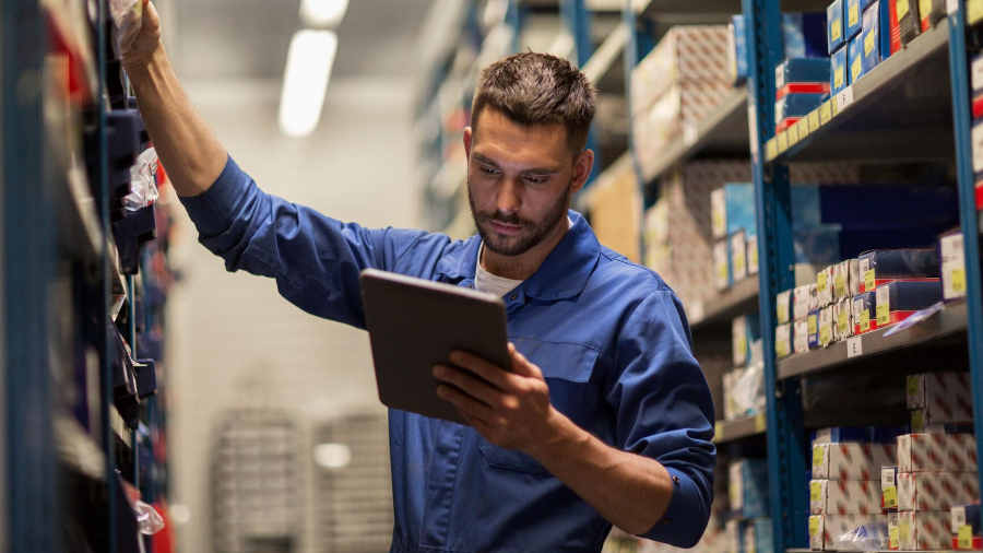 A man in a blue work uniform stands in a warehouse aisle, holding a tablet in one hand and reaching for a box on a shelf with the other. Shelves around him are filled with various small boxes and items.