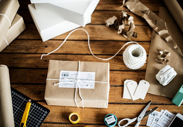 A neatly wrapped package with a shipping label sits on a wooden table surrounded by packing supplies, including white twine, scissors, shipping labels, blank tags, a utility knife, tape, and sheets of kraft paper.