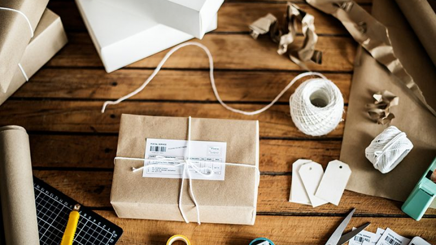 A neatly wrapped package with a shipping label sits on a wooden table surrounded by packing supplies, including white twine, scissors, shipping labels, blank tags, a utility knife, tape, and sheets of kraft paper.