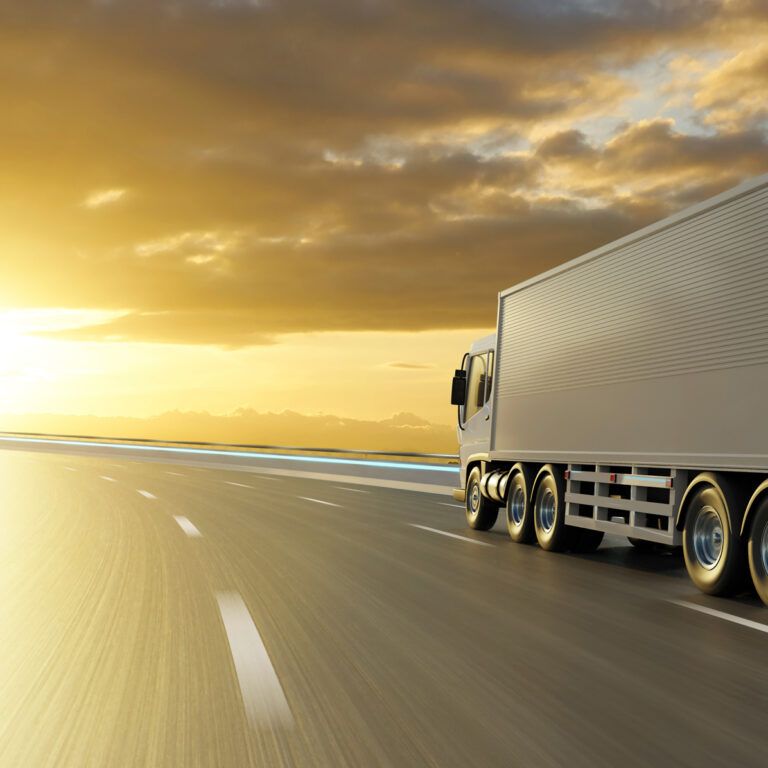 A large white truck drives along a curved highway at sunset, with dramatic clouds and golden sunlight illuminating the road and mountains in the distance.