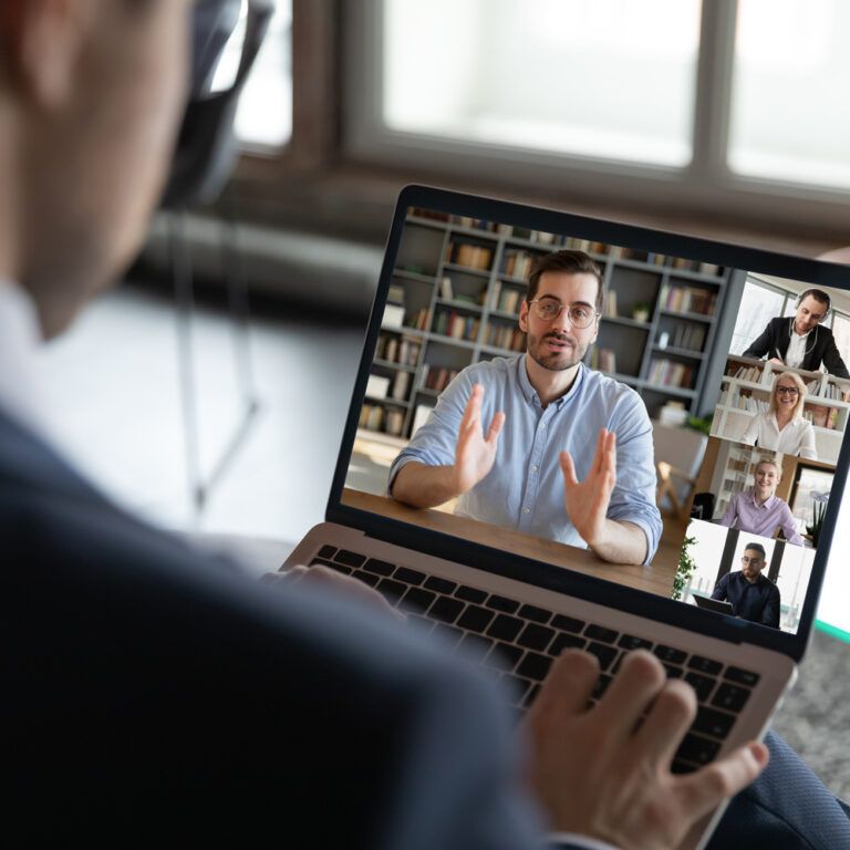 A person in a suit participates in a video conference on a laptop, with five people visible on the screen, each in their own window, engaged in a discussion. A potted plant and window are in the background.