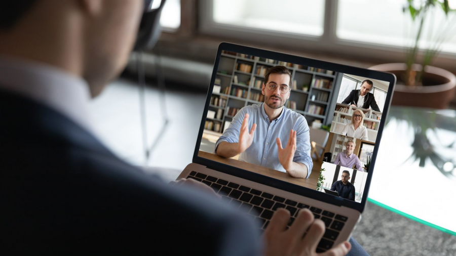 A person in a suit participates in a video conference on a laptop, with five people visible on the screen, each in their own window, engaged in a discussion. A potted plant and window are in the background.
