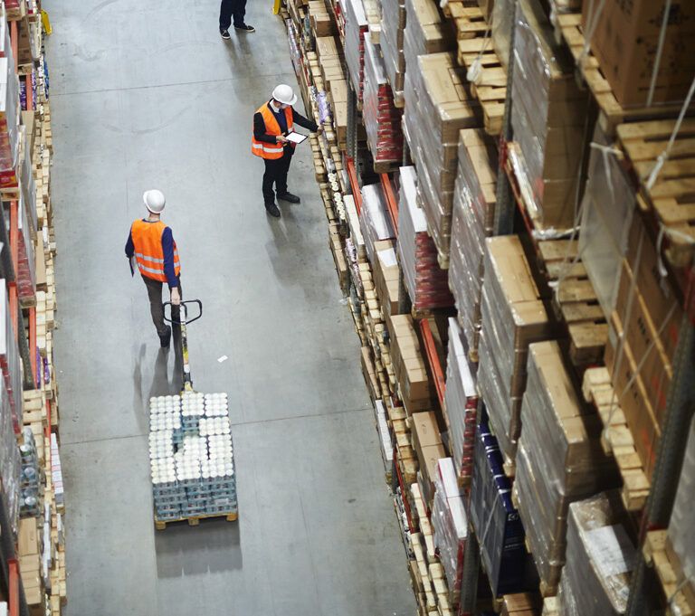 High-angle view of two workers in orange safety vests and white helmets moving and inspecting goods in a large warehouse filled with tall shelves stacked with boxes and pallets.