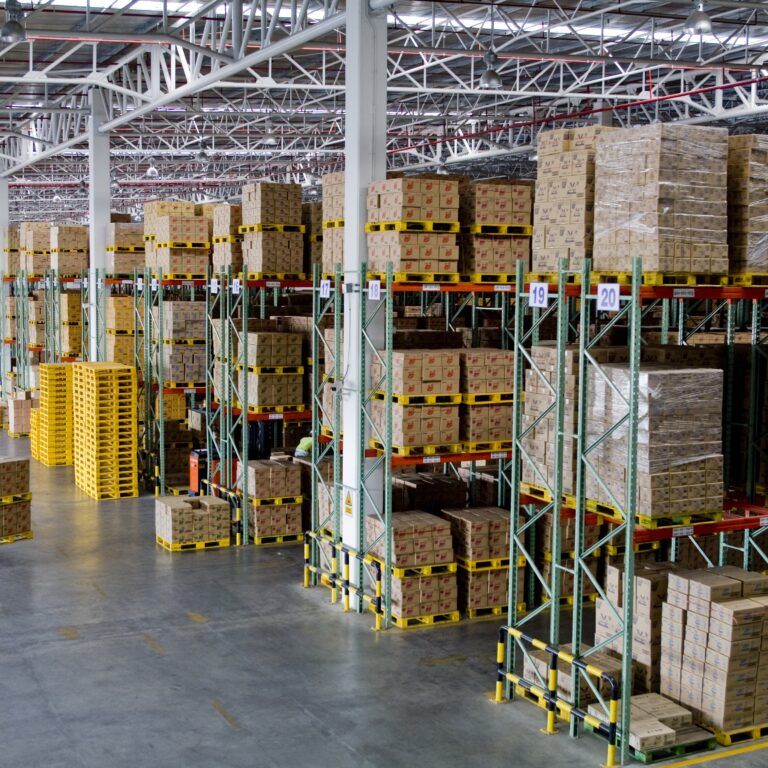 A spacious, well-lit warehouse with high shelves stacked with cardboard boxes, organized racks of goods in rows, and a worker operating a forklift—motivated by incentive based pay—on the left side of the image.
