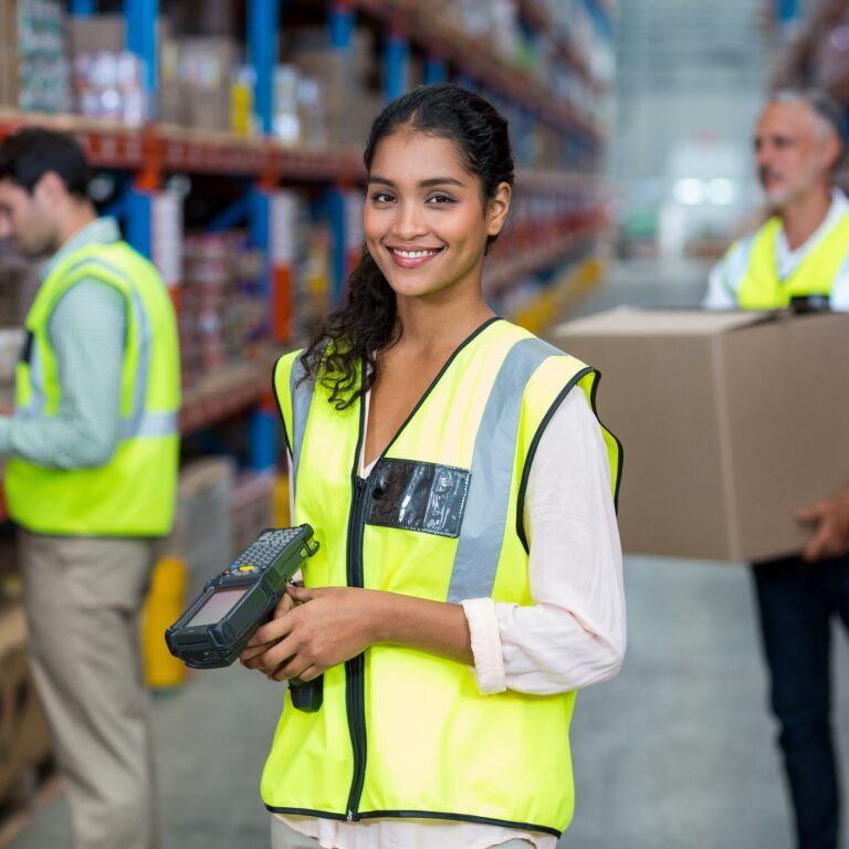 A woman in a yellow safety vest smiles while holding a barcode scanner in a warehouse; two coworkers in safety vests work in the background among shelves of boxes.